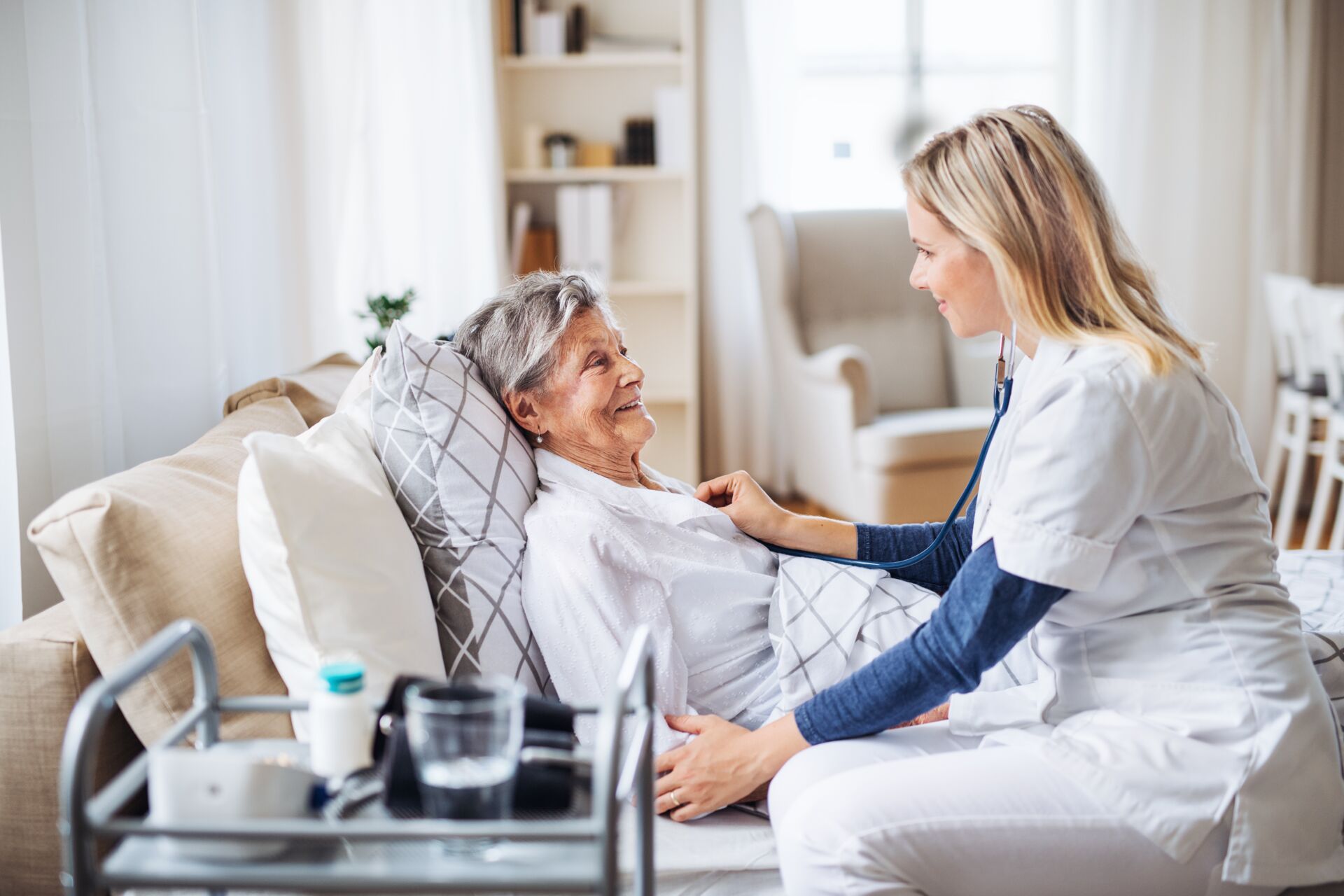 Eine Krankenschwester sitzt bei einer älteren Dame am Pflegebett und misst Blutdruck