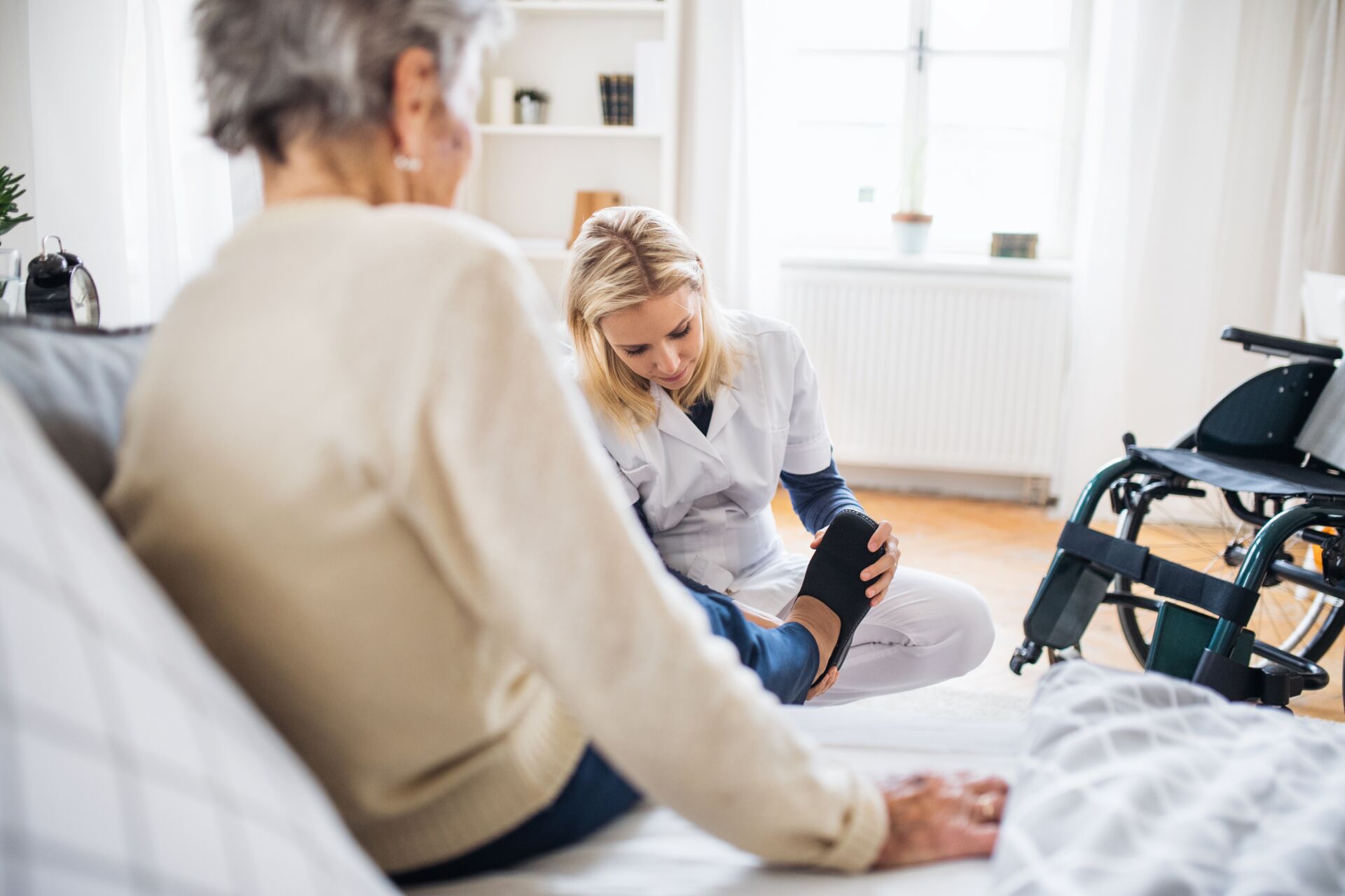 Häuslicher Intensivpflegedienst & Alltagsbegleitung Eine blonde Pflegekraft hilft einer älteren Frau, die auf einem Sofa sitzt beim Schuhe anziehen. Im Hintergrund steht ein Rollstuhl.