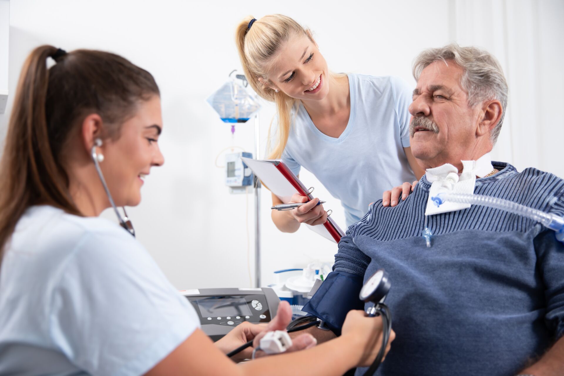 Ambulante Intensivpflege & Heimbeatmung Eine junge Krankenschwester misst den Blutdruck eines älteren Herrn, während eine zweite Krankenschwester lächelnd daneben steht.
